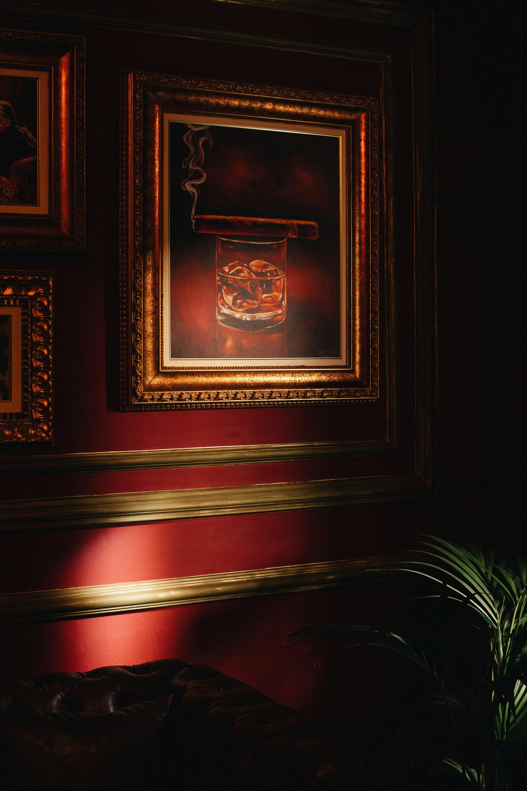 A sophisticated painting of whisky and cigar in a golden frame, illuminated on a rich red wall.