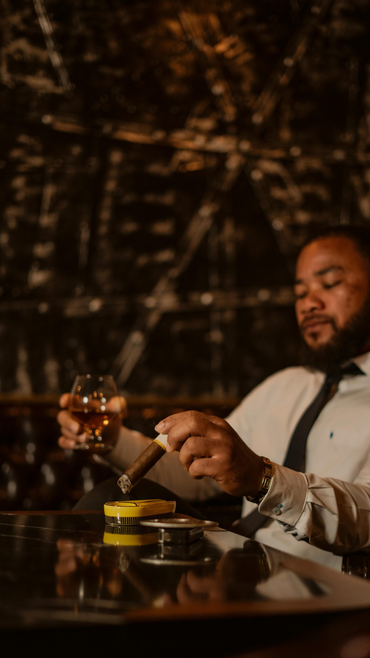 A well-dressed man relaxes with a cigar and drink in a sophisticated indoor setting.